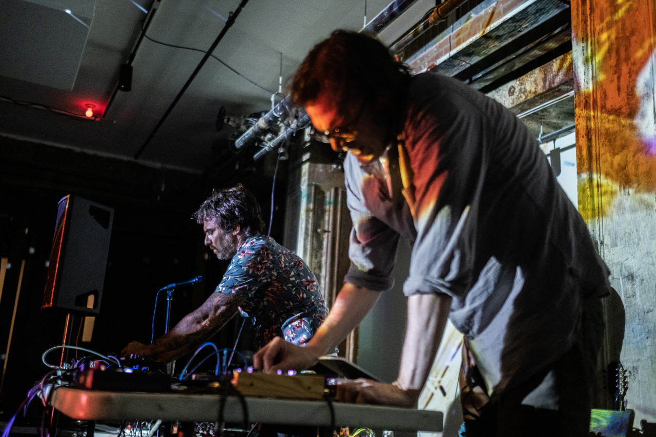 Two performers leaning over tables use electronic instruments. The performer in the foreground and a column behind them is bathed in orange light. A single red colored light bulb is visble on the ceiling.