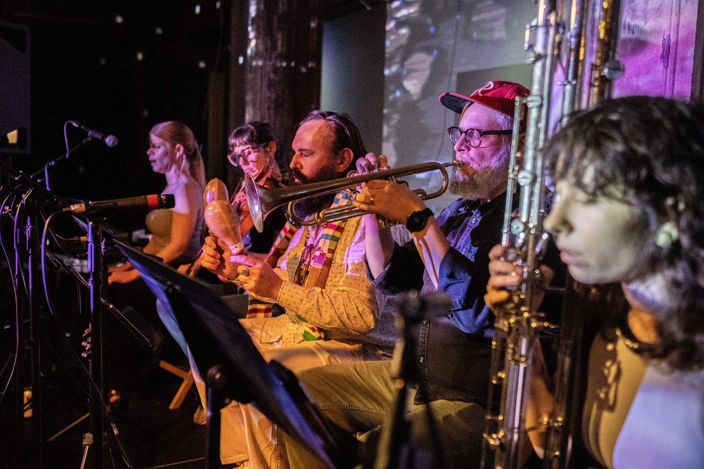 Five performers sitting in a row playing various wind, string, and percussion instruments. They are facing microphone and sheet music stands. There is a pink-tint covering the musicians and the wall behind them.