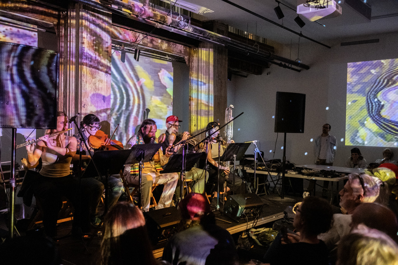 Five performers sitting in a row playing various wind, string, and percussion instruments. They are facing microphone and sheet music stands. Multicolored patterned projections and disco ball reflections cover the two walls and musicians. Audience members are visible around the stage.