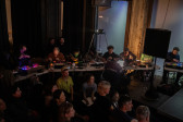 Eight musicians sitting side by side at tables use electronic instruments. Various colored lights emit from each audio board. There are audience members in front of and behind the musicians. The room is dimly lit with a strip of blue light on a wall.