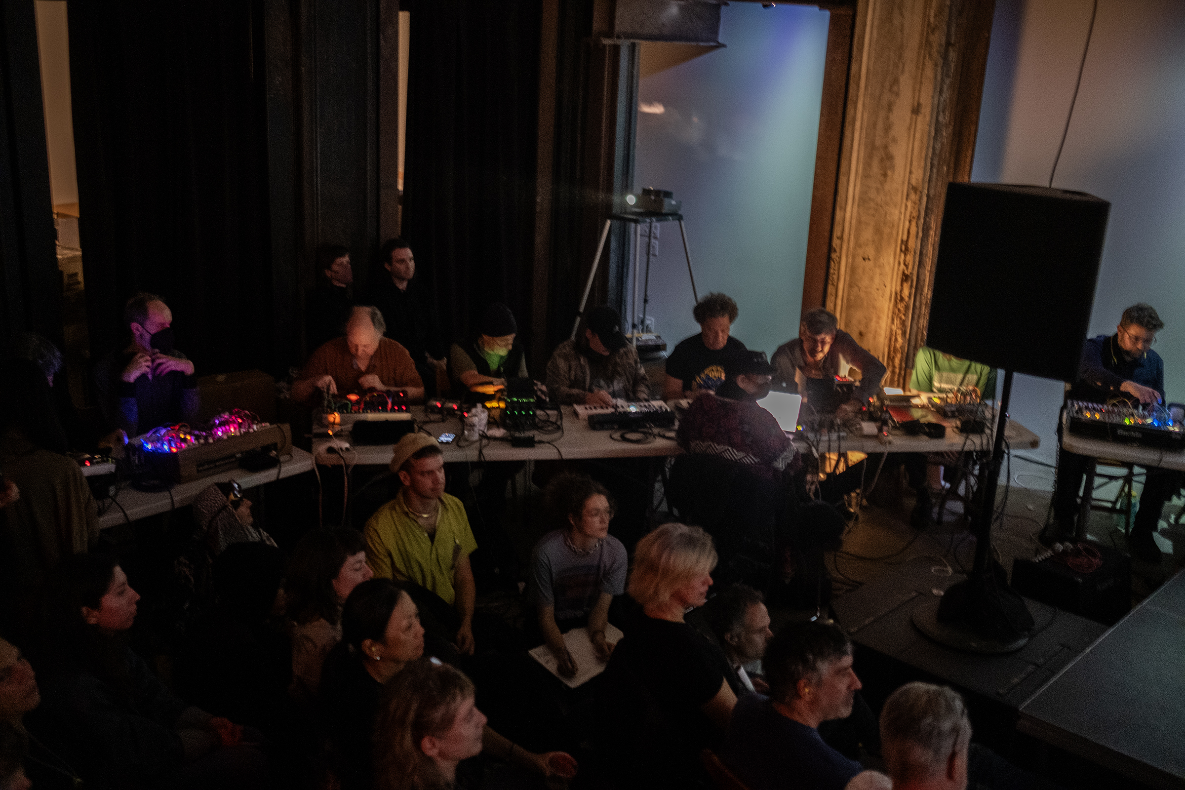 Eight musicians sitting side by side at tables use electronic instruments. Various colored lights emit from each audio board. There are audience members in front of and behind the musicians. The room is dimly lit with a strip of blue light on a wall.