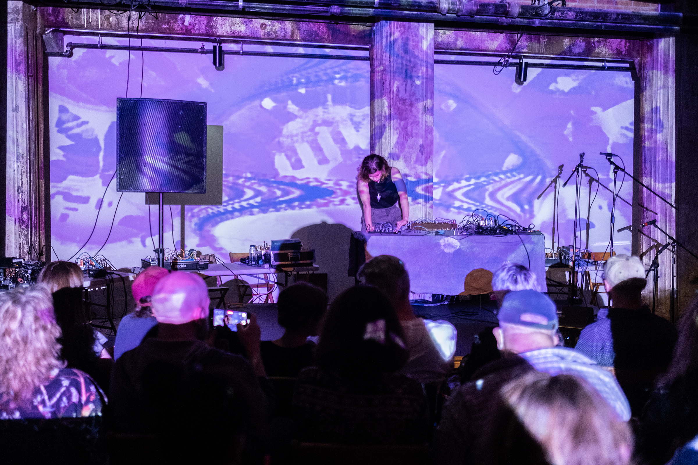 Solo performer on stage behind a table of electronic instruments with a large speaker to their right. The walls and musician are covered with pink and purple patterned projections. A group of audience members are visible in the foreground.