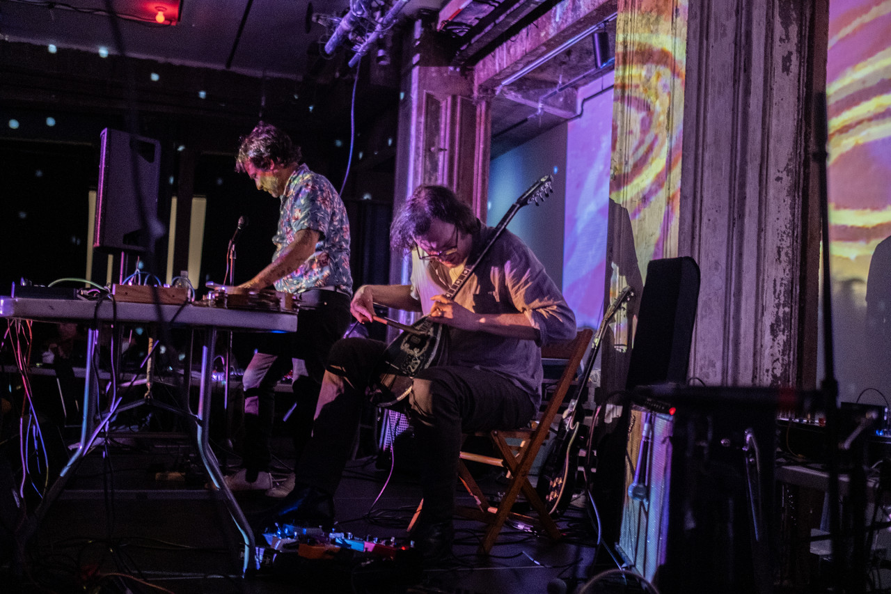 Two musicians on stage use various instruments. The musician in the foreground plays a mandolin. The musician behind them is using electronic instruments on a table. There are yellow and pink patterned projections, a red light bulb, and reflections from a disco ball around the room.