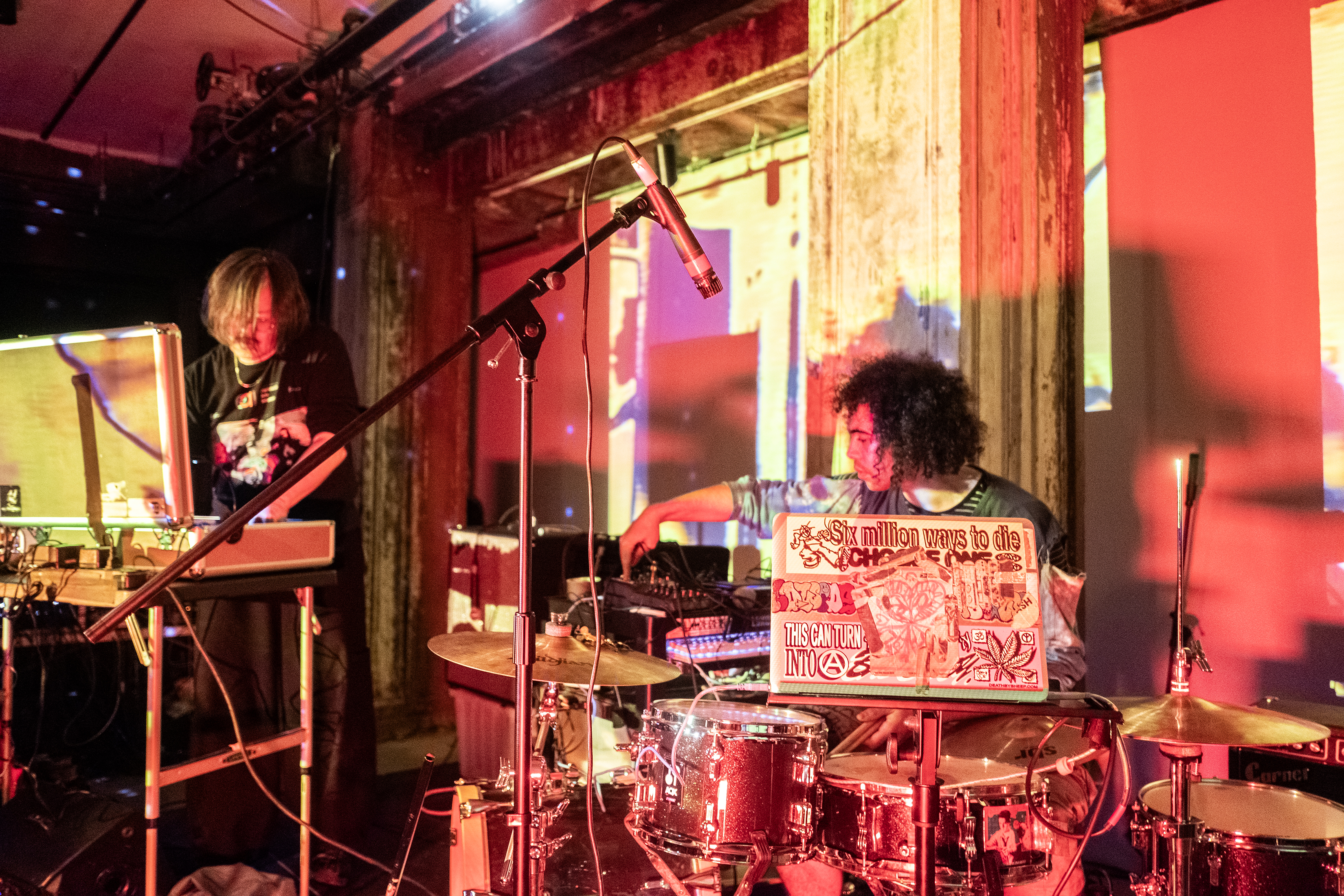 Two performers on stage, the one on the right is playing the drums and using electronic audio equipment, and the performer on the left is leaning over a large case on top of a table. The room is bathed in yellow, pink, red, and purple light. The wall behind them is projected with abstract patterns.