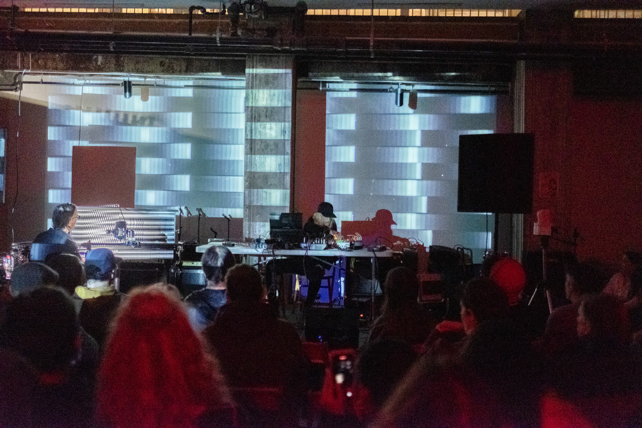 A crowd faces a stage where a musician is sitting over various electronic instruments and audio equipment. The audience members are bathed in red light. The wall behind the musician is projected with monochromatic gradients.
