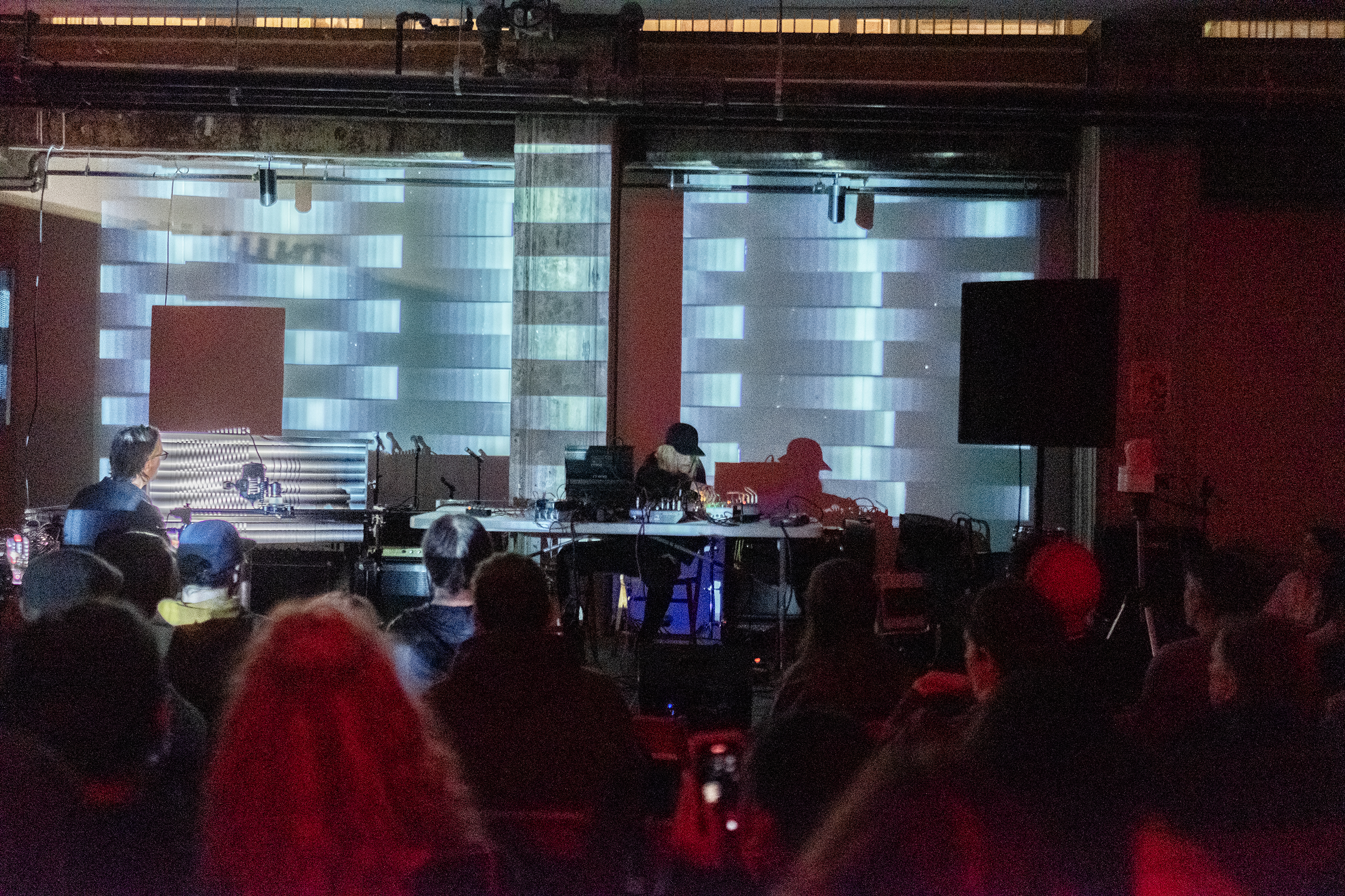 A crowd faces a stage where a musician is sitting over various electronic instruments and audio equipment. The audience members are bathed in red light. The wall behind the musician is projected with monochromatic gradients.