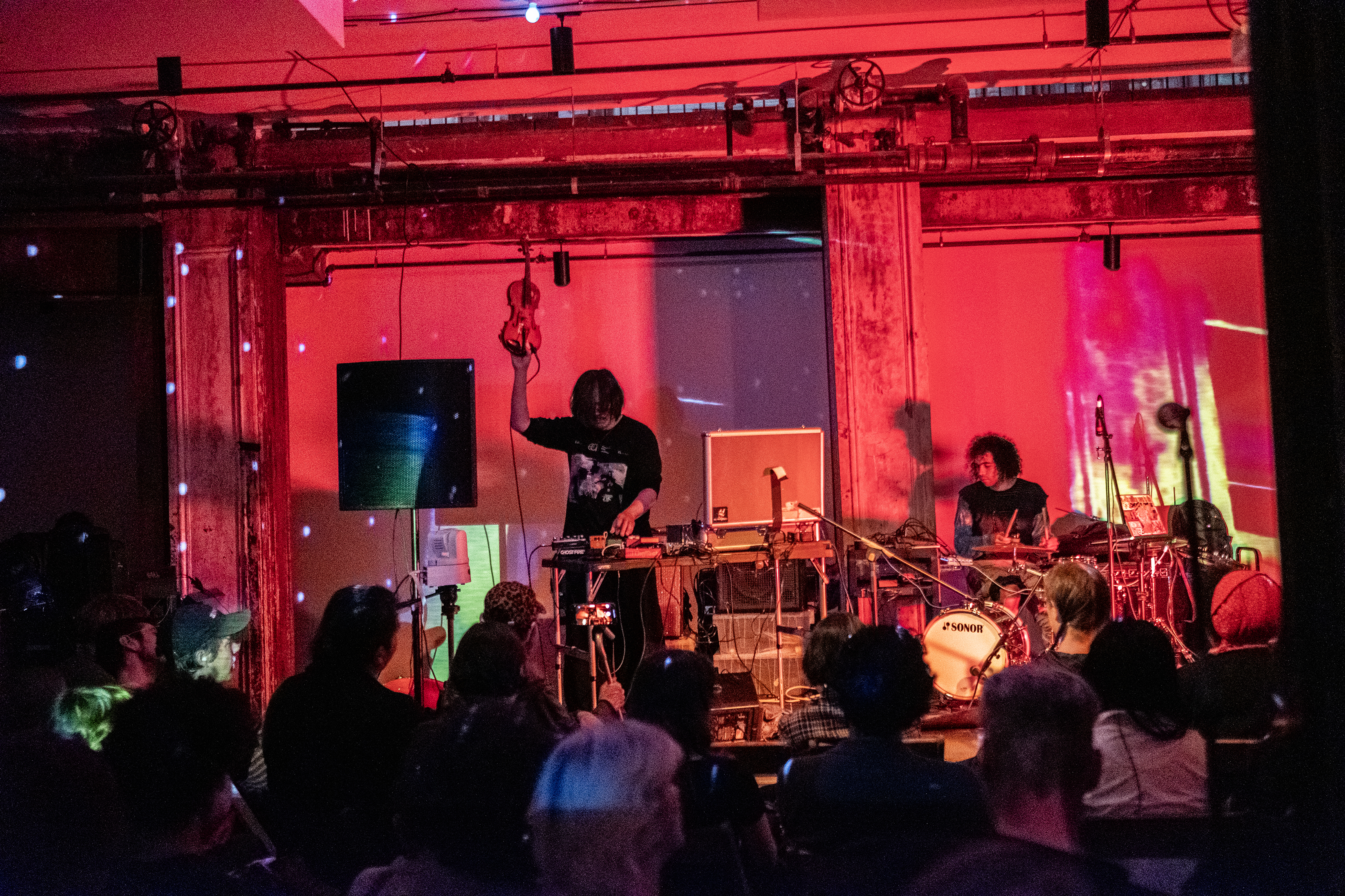 Two performers on stage in front of a seated audience. The musician on the left is holding a violin in the air as they use audio equipment on the table in front of them. The musician on the right is sitting playing the drums. The room is red-tinted, and white dots reflected from a disco ball cover the walls.
