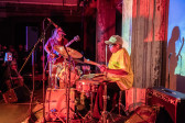 Image of a performer on stage in a yellow shirt and baseball cap playing the drums. They are facing an audience. To their right is another performer playing the guitar. The room is dark with red and blue-tinted light.
