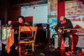 Two musicians sit on stage. The musician on the left is leaning over a table and using electronic instruments. The musician on the right is looking down and playing the guitar. The stage is lit with red and white lights and projected with a patchworks of patterns.