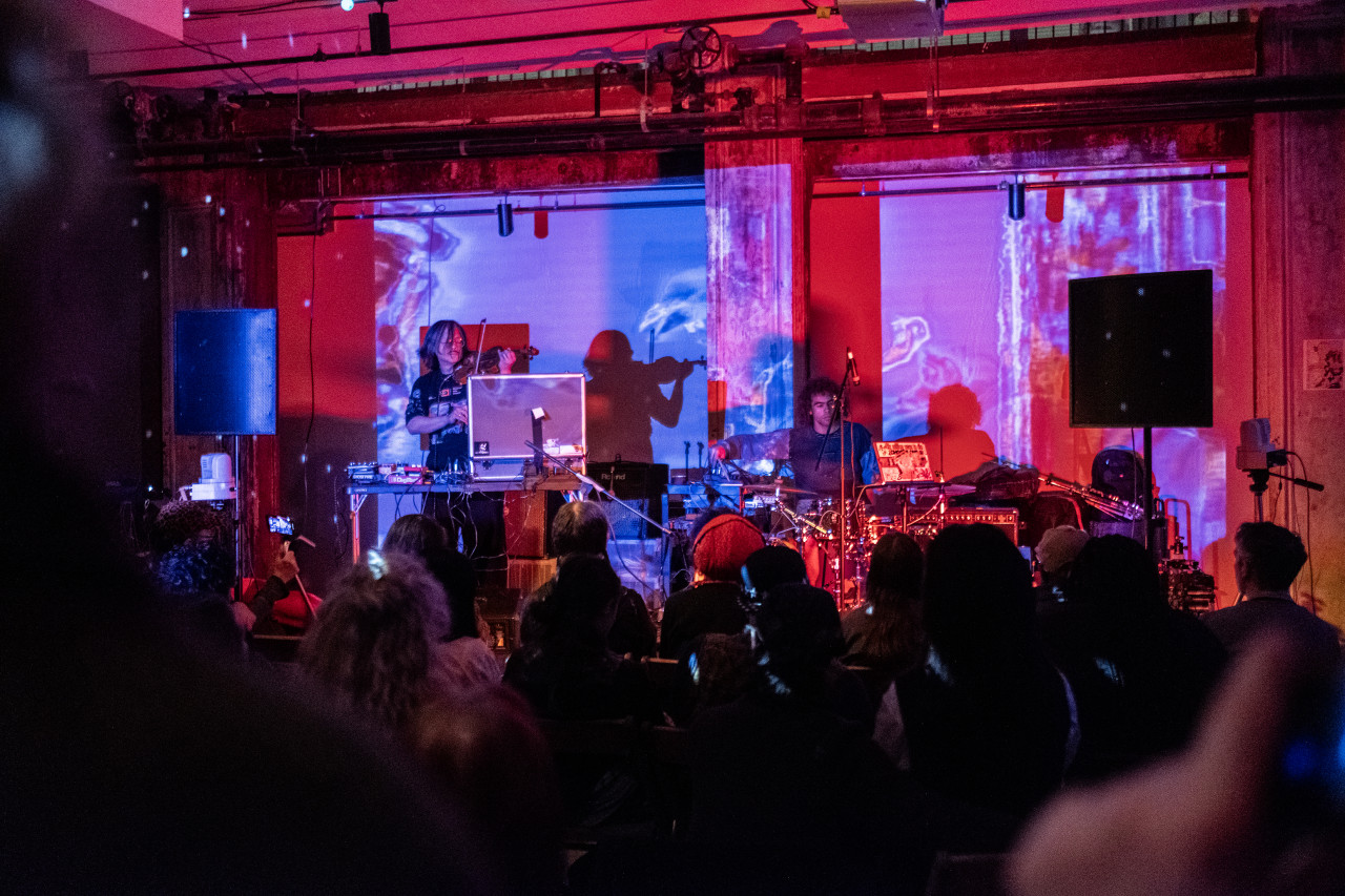 Two performers on stage in front of a seated audience. The musician on the left is playing the violin, and the musician on the right is playing the drums. The room is bathed in red and blue light. The wall behind the performers is projected with blue and purple graphics.