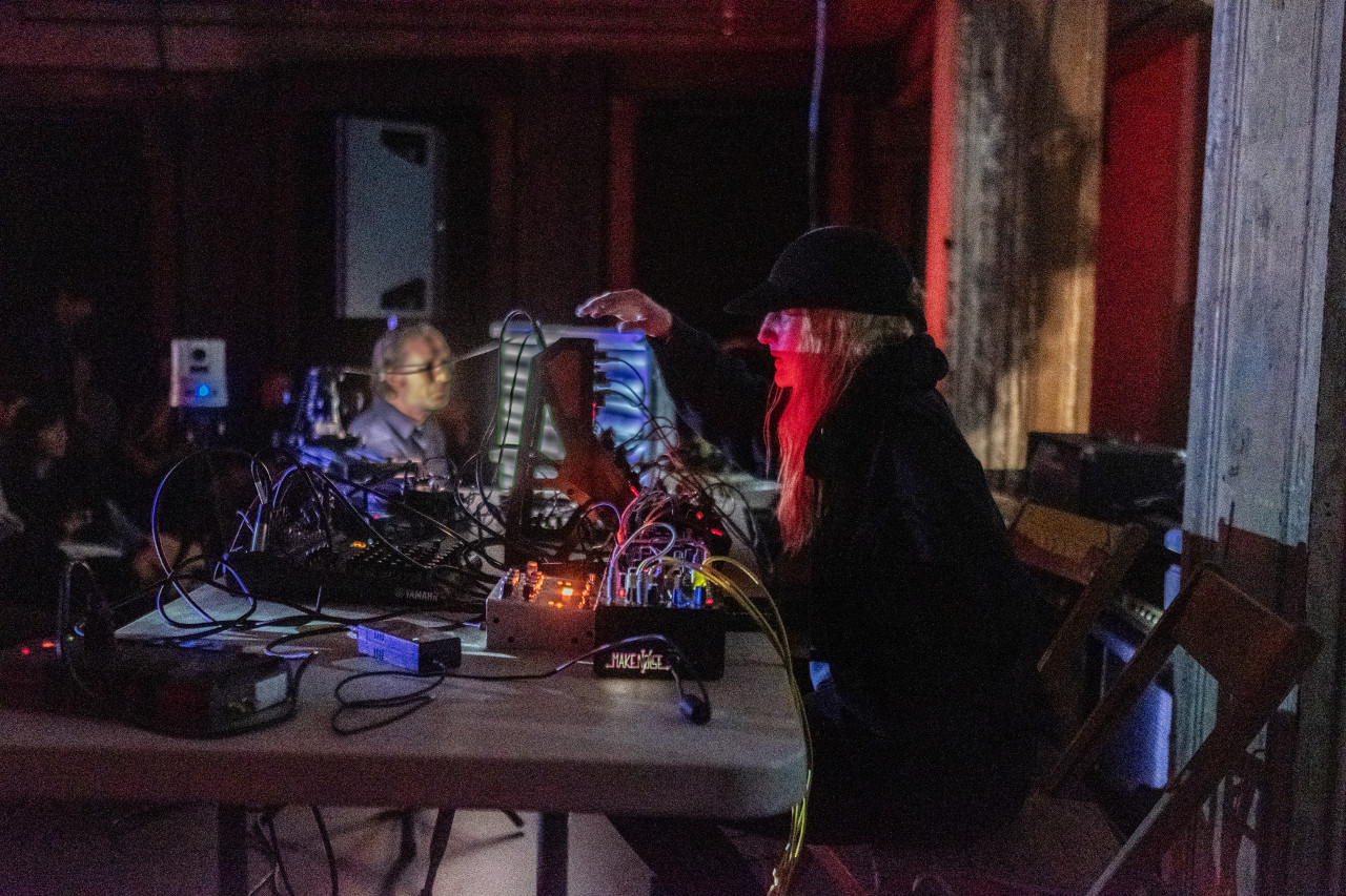 A performer with long blonde hair and a black baseball cap sits on stage at a table covered in electronic instruments. Their face is lit with red light. The background is dark, but a few audience members are visible.