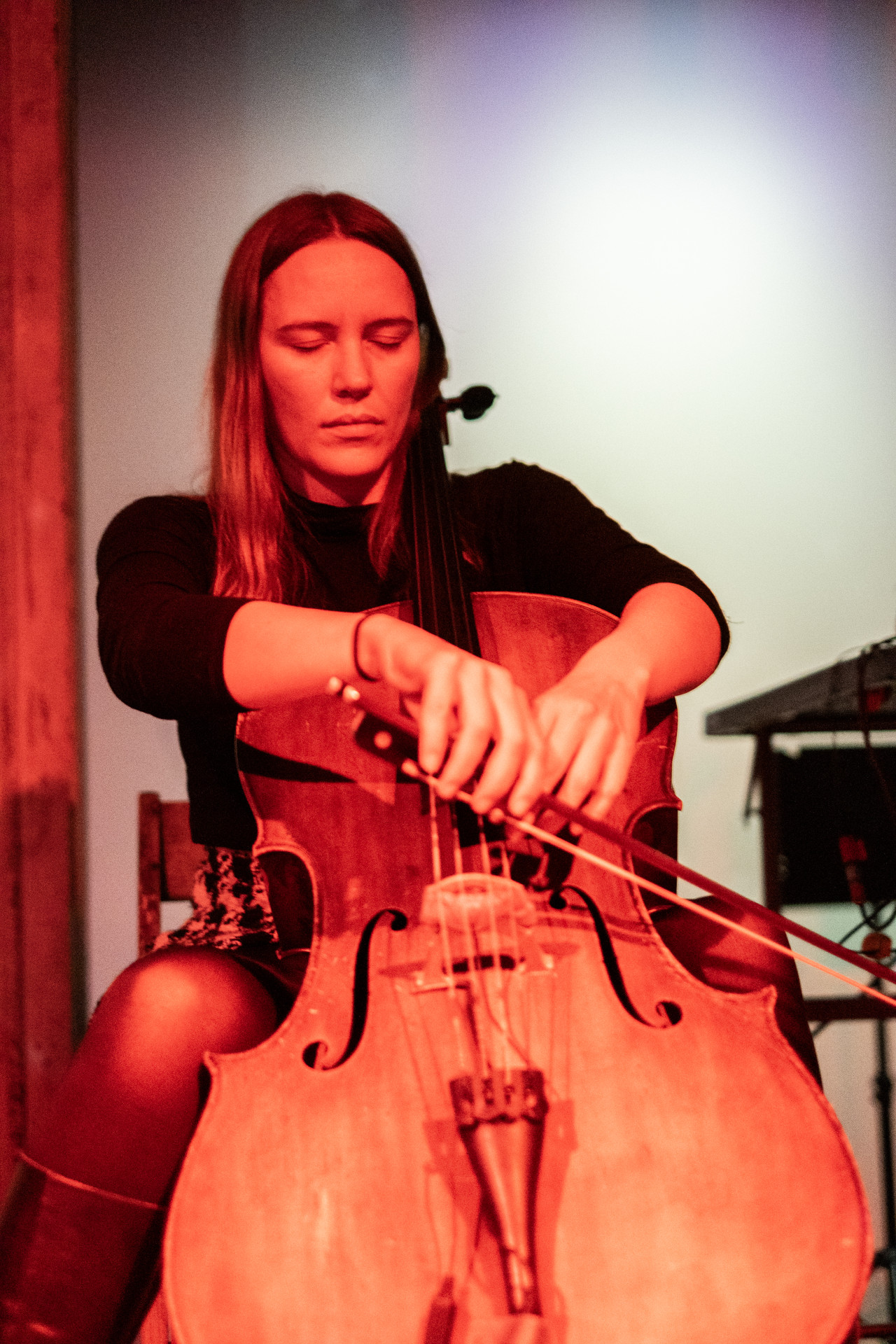 A performer sitting down with their eyes closed playing the cello. They are using one hand to play with a bow and the other hand is on the strings. They are tinted red from the lighting, the back wall is lit with white and purple light.