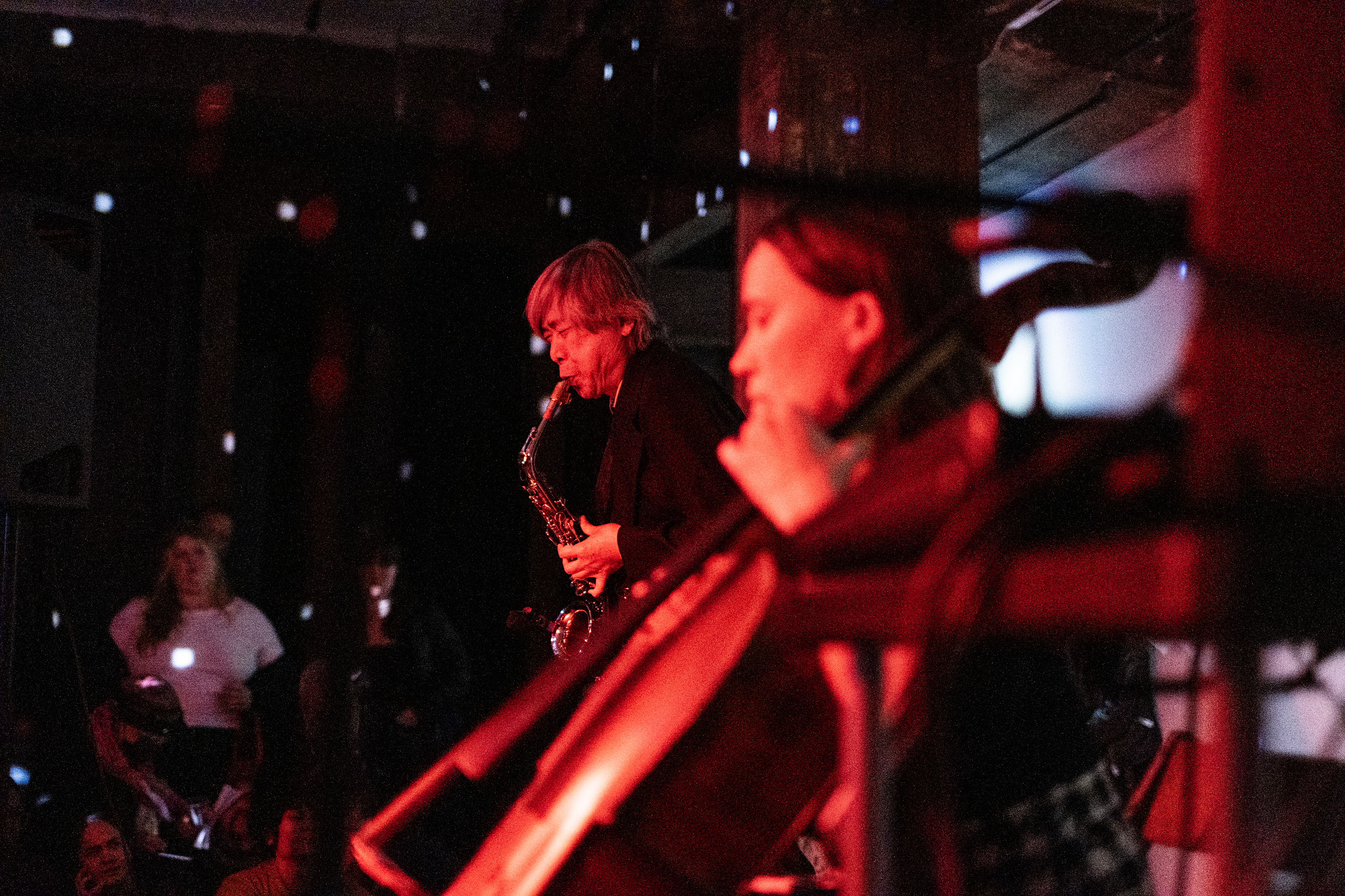 A view from the side of the stage of two performers. The performer in the back is playing the saxophone and has their eyes closed. The musician in the foreground is out of focus. They are playing the cello. Both musicians are tinted red, as well as the audience on the left side, and the room has white dots of light reflecting from a disco ball.
