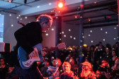 A performer playing a blue retro-style guitar stands in front of a large audience. The crowd faces the musician, some of them are sitting closer to the stage and others stand in the background. A red stage light on the ceiling illuminates the room. White dots reflect around the room from a disco ball. 