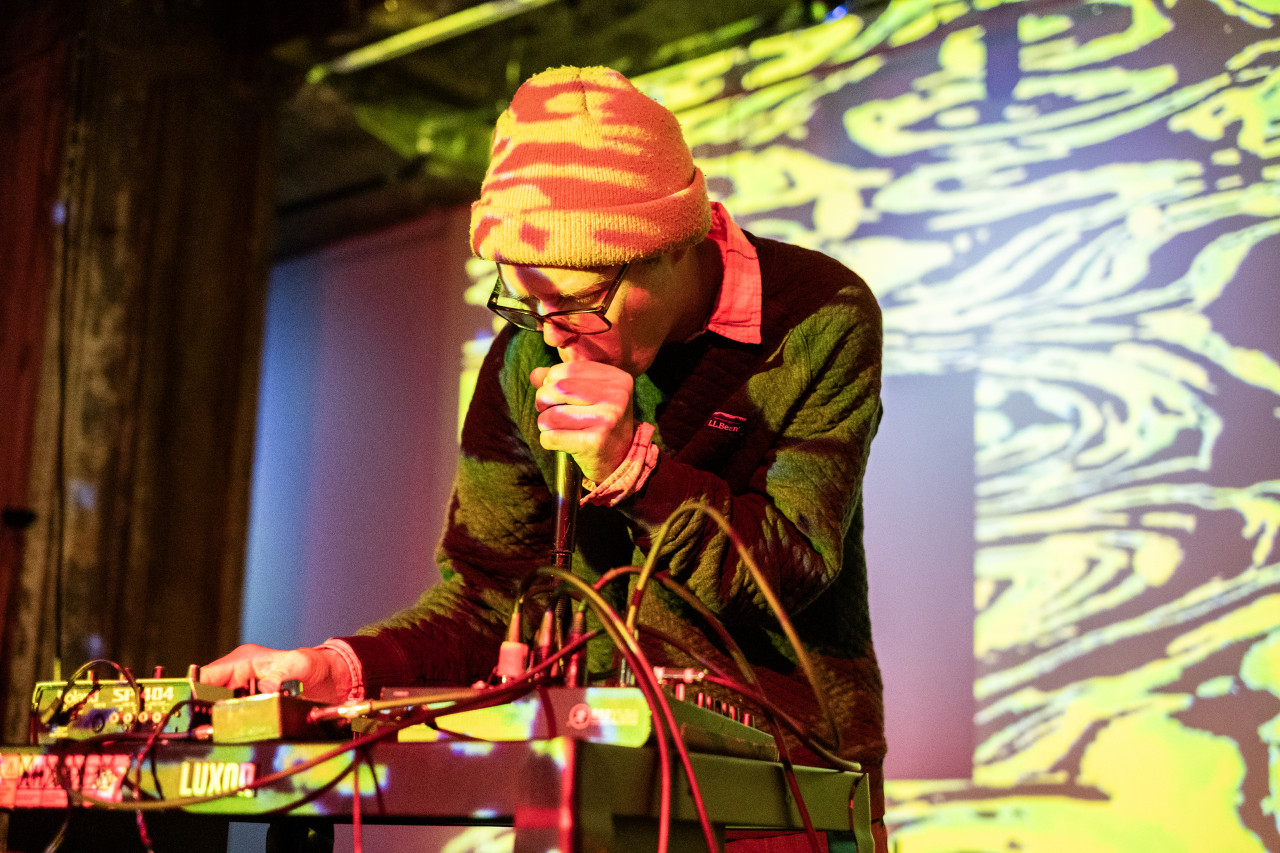 A performer on stage leaning over a table with electronic instruments, holding a mic close to their mouth. They are bathed in yellow abstract projections, which also cover the wall.