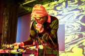 A performer on stage leaning over a table with electronic instruments, holding a mic close to their mouth. They are bathed in yellow abstract projections, which also cover the wall.
