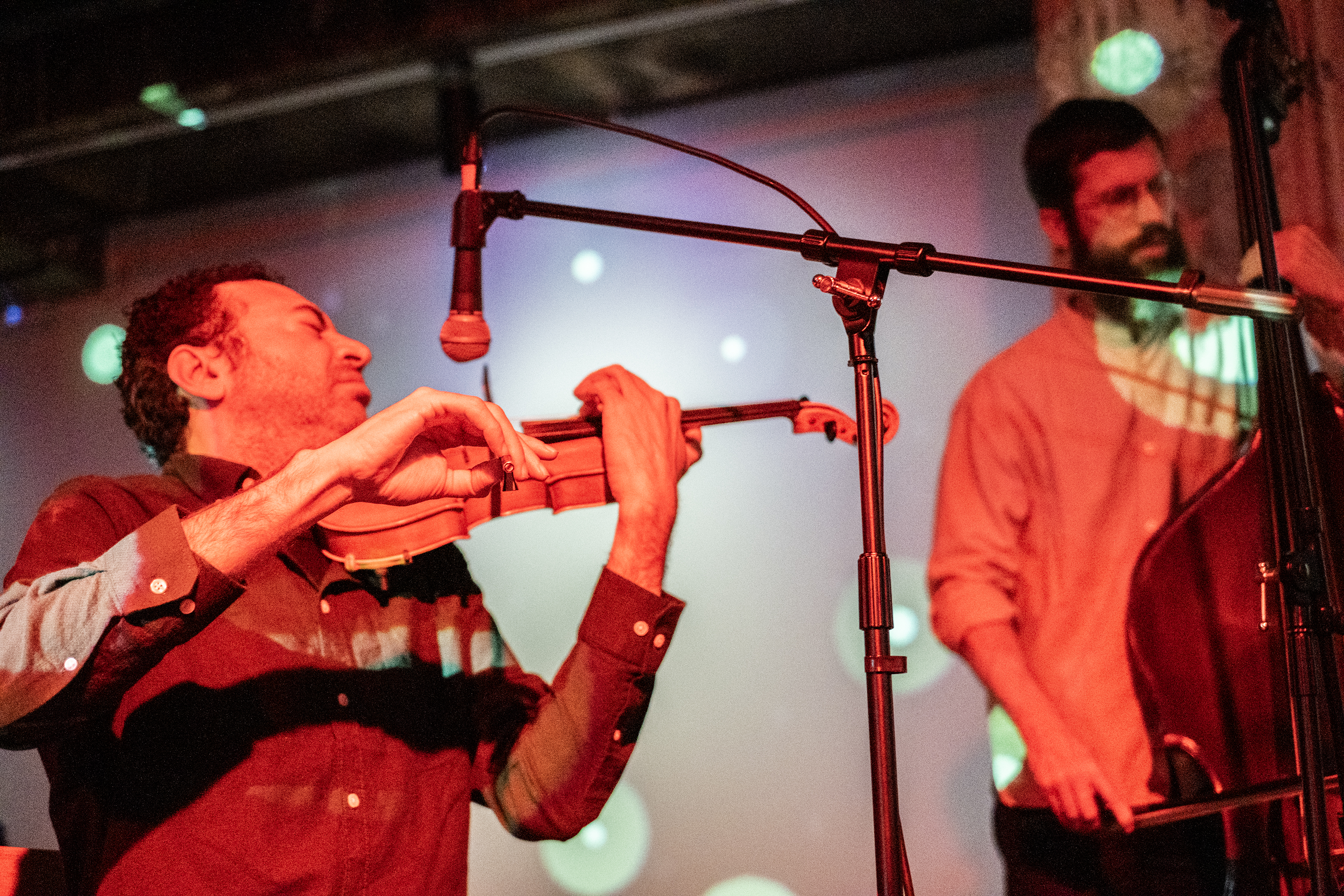 A close view of a performer playing the violin. Their hand using the bow is in focus and the rest of the image is slightly blurred. They are playing into a microphone on a stand. To their left a performer plays the upright bass. Both musicians are covered with red light and images of projected green circles.