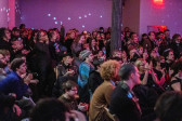 A room full of audience members in a pink-tinted room. The people in the front are sitting, and the ones behind are standing close together. Many people are clapping. The room is filled with patterns of white dots reflecting from a disco ball.
