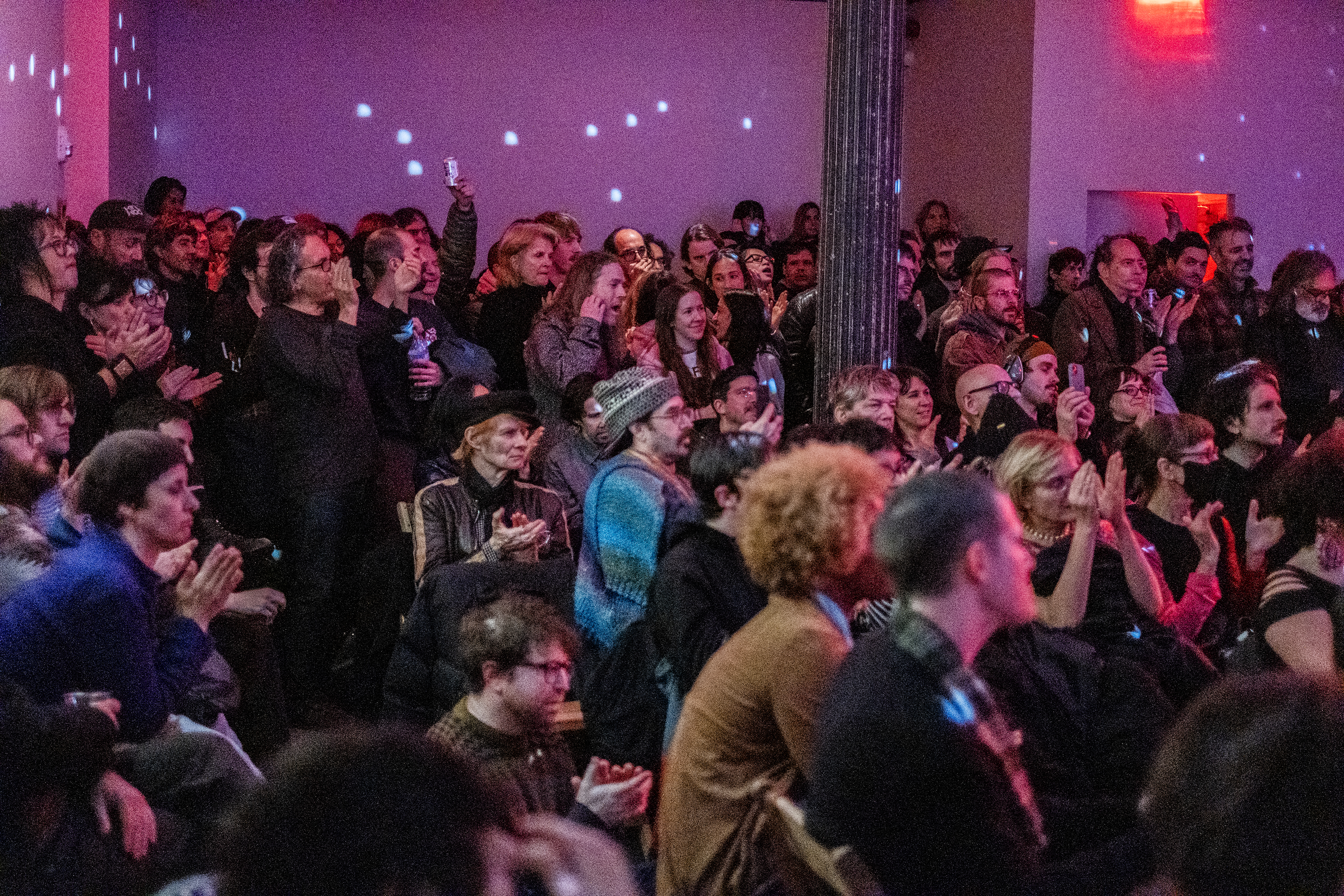 A room full of audience members in a pink-tinted room. The people in the front are sitting, and the ones behind are standing close together. Many people are clapping. The room is filled with patterns of white dots reflecting from a disco ball.