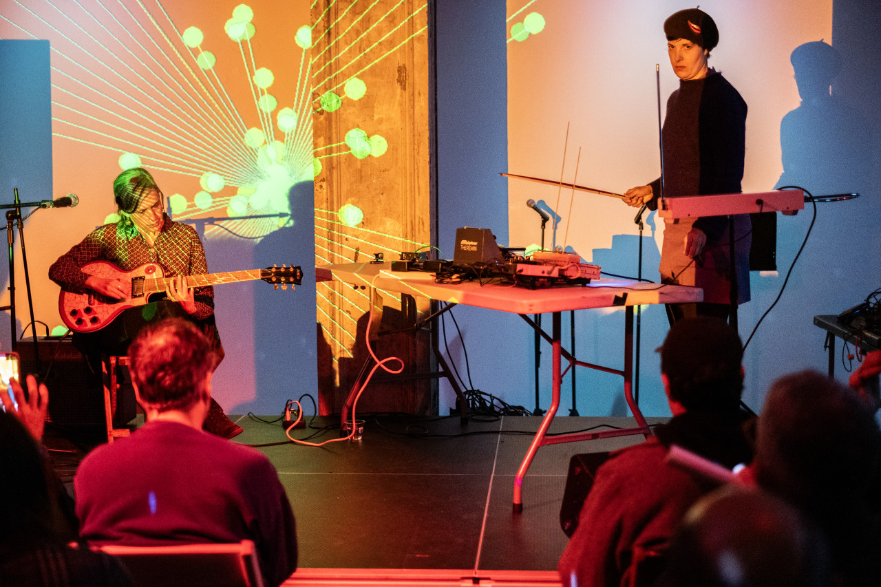 Two performers on stage with a few audience members in the foreground. The performer on the left is sitting, looking down, and playing guitar. The performer on the right is standing and holding a bow. The audience and musicians are tinted with orange and red toned lights. An abstract orange and green projection covers the back wall.