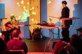 Two performers on stage with a few audience members in the foreground. The performer on the left is sitting, looking down, and playing guitar. The performer on the right is standing and holding a bow. The audience and musicians are tinted with orange and red toned lights. An abstract orange and green projection covers the back wall.