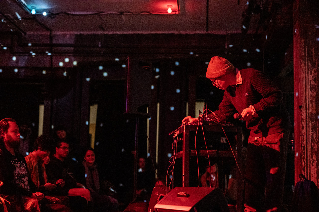 A performer facing an audience in a dim, red-tinted room. The musician is standing over a table covered in electronic instruments. Patterns of white dots reflecting from a disco ball surround the room.