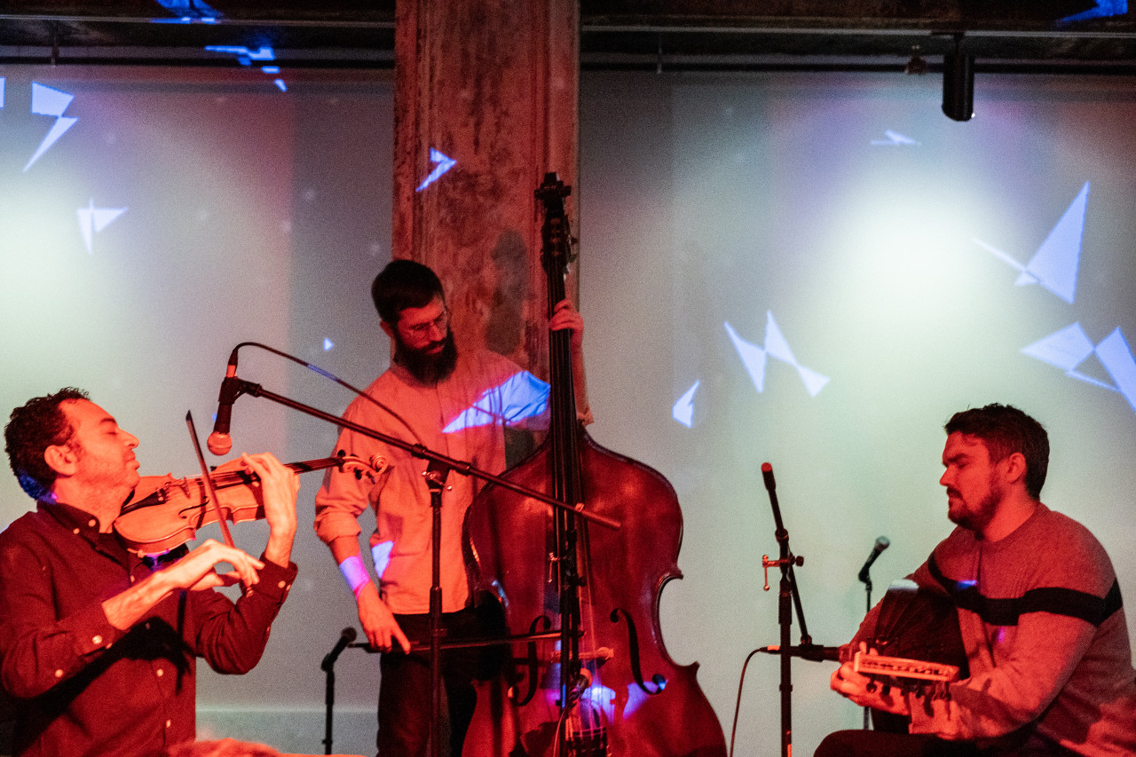 An image of three performers on stage facing each other. The musician in the middle is standing and playing an upright bass with a bow. On their right side a musician is sitting and playing violin. On the other side a musician is sitting playing an instrument similar to a mandolin. They are all bathed in red light and projections of white angular shapes cover the musicians and the wall.