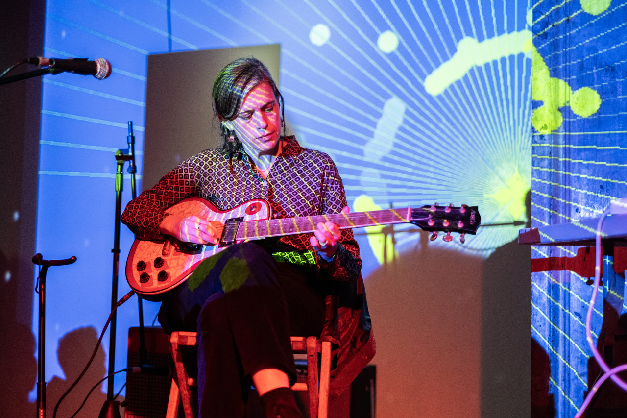 A performer sitting down playing the guitar is bathed in colorful light. They are wearing a geometric print shirt and a blue, green, and white projected pattern covers their face and the wall behind. There are two mic stands to their right.