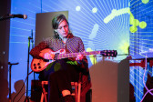A performer sitting down playing the guitar is bathed in colorful light. They are wearing a geometric print shirt and a blue, green, and white projected pattern covers their face and the wall behind. There are two mic stands to their right.