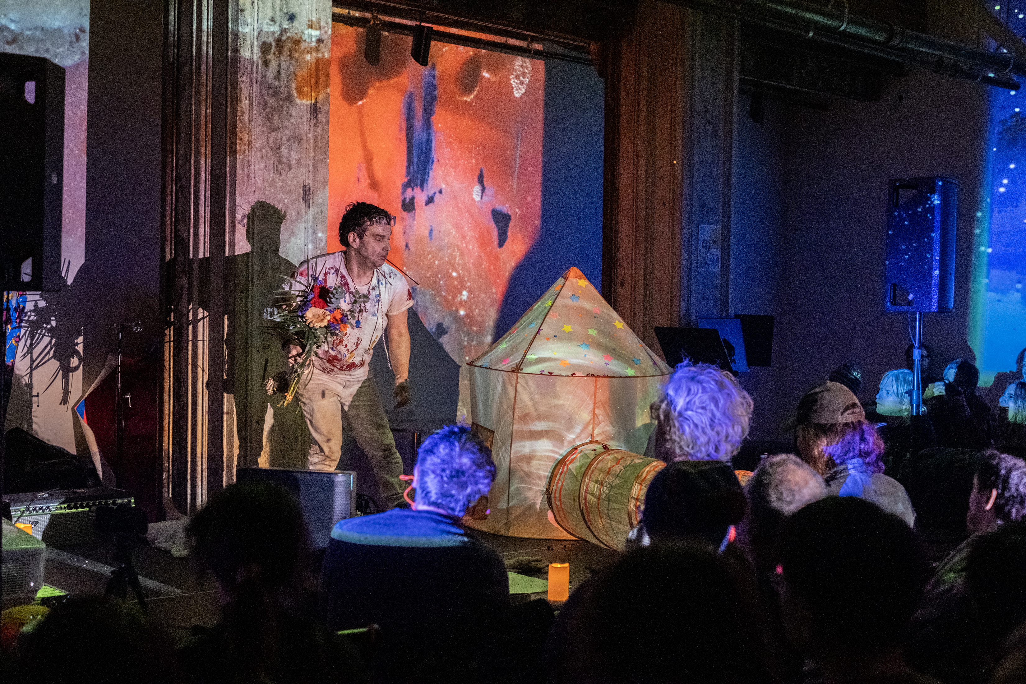 Color image of a performer and a small white tent on a stage. The performer is on the left, wearing all white, with a bouquet of flowers in their hand and a stick of insense in their mouth. On the right side of the stage, the tent is lit up with multicolored projections. The top of the tent is decorated with colorful stars. The walls in the background, as well as the audience in the foreground, are lit by patterned projections and blue lights.