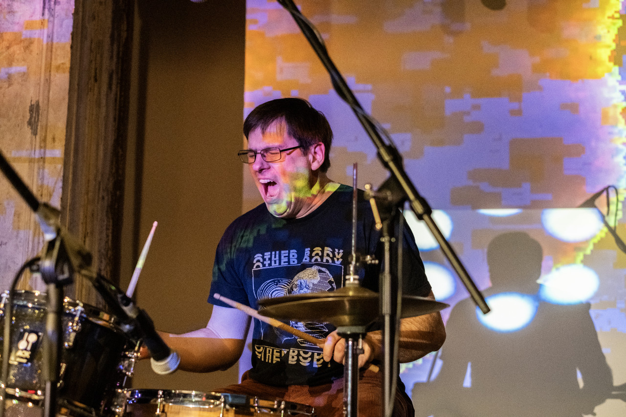 Color image of a performer playing the drums. Their eyes are closed and their mouth is open, they are wearing a black t-shirt and glasses. The face of the performer and the wall in the background are decorated with colorful pattern projections.