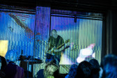 Color image of a performer standing in the center of the frame, looking down while playing an electric guitar. In the foreground the backs of audience members are blurred. The scene is decorated with an abstract striped projection resembling a screen glitch.