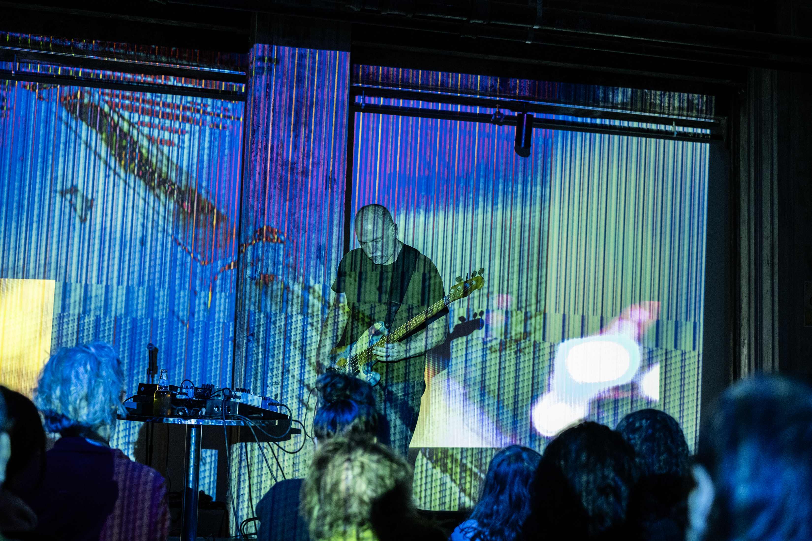 Color image of a performer standing in the center of the frame, looking down while playing an electric guitar. In the foreground the backs of audience members are blurred. The scene is decorated with an abstract striped projection resembling a screen glitch.