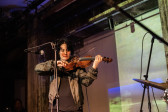 Color image of a performer standing up playing the violin. They are standing in front of a mic stand with their eyes closed. A drum kit is barely visible in the right corner of the image. The wall in the background is tinted with green and blue lights.