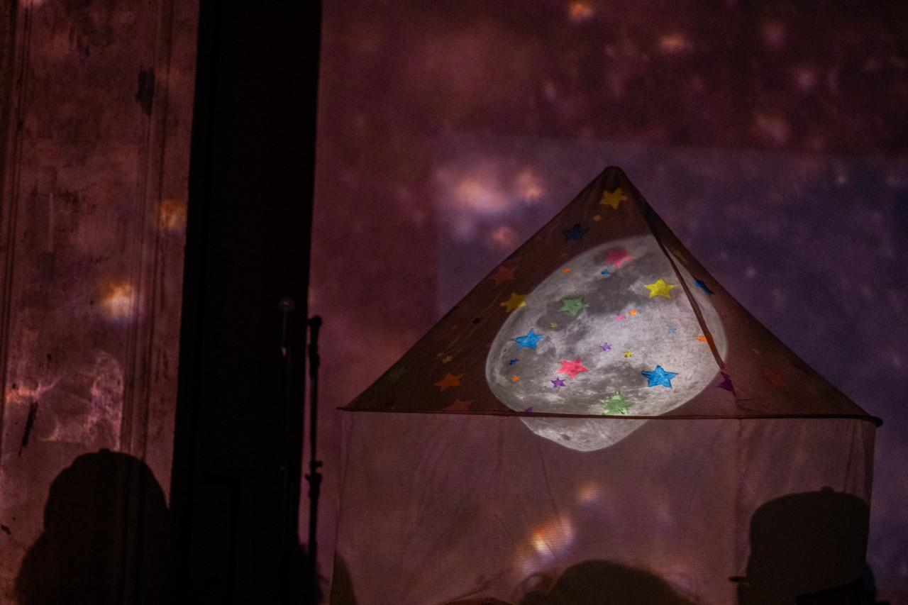 Color image of a fabric tent with an image of the moon projected on the top. The top of the tent is decorated with multicolored stars. The tent and the wall behind it are bathed in pale pink light, with small speckles of white light.