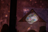 Color image of a fabric tent with an image of the moon projected on the top. The top of the tent is decorated with multicolored stars. The tent and the wall behind it are bathed in pale pink light, with small speckles of white light.