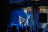 Color image of a performer on stage playing the electric guitar. A couple rows of audience members are visible in the bottom edge, with their backs facing the camera. The performer and the wall behind them are bathed in blue light. An image resembling a galaxy is projected on the wall.