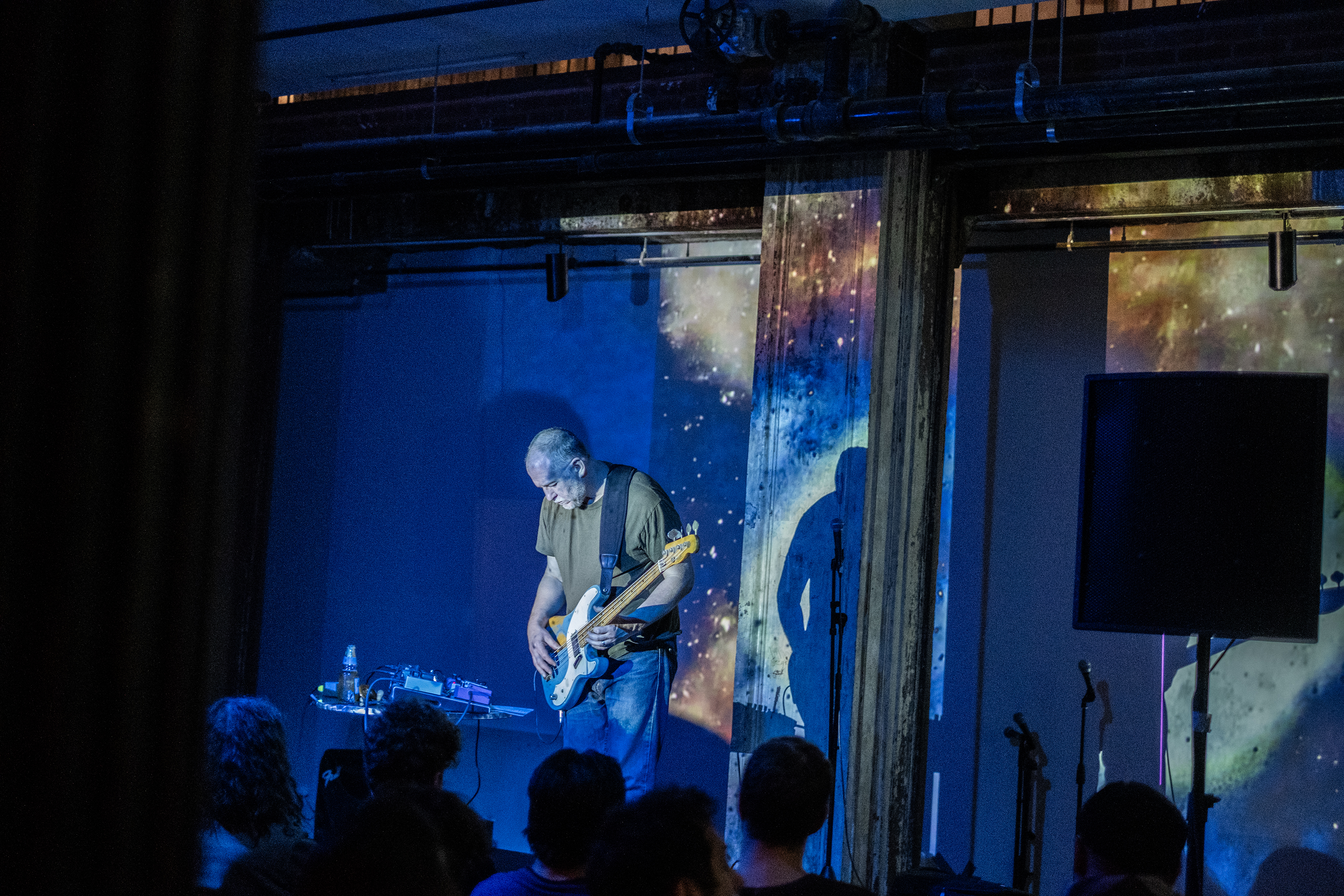 Color image of a performer on stage playing the electric guitar. A couple rows of audience members are visible in the bottom edge, with their backs facing the camera. The performer and the wall behind them are bathed in blue light. An image resembling a galaxy is projected on the wall.