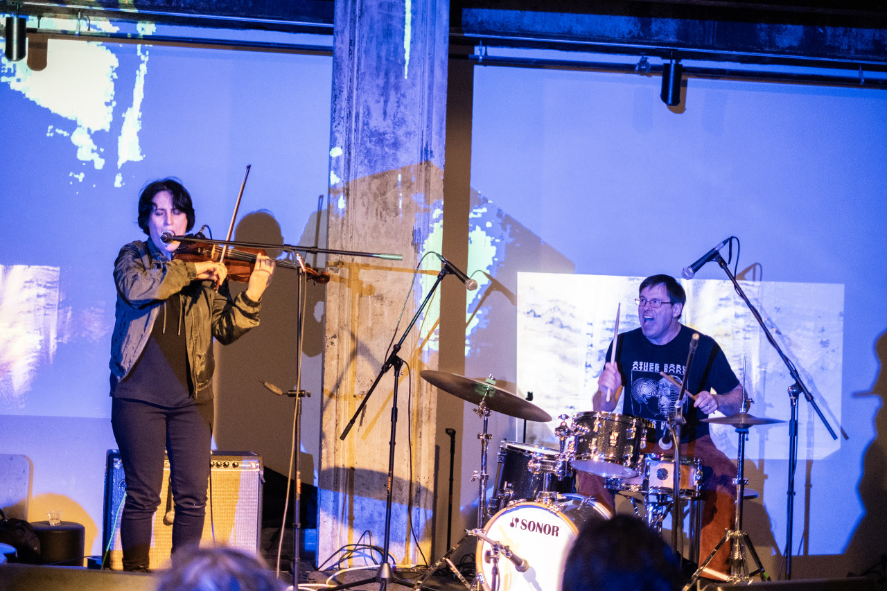 Color image of two performers on stage. The performer on the left is standing as they sing into a microphone and play the violin. The performer on the left is sitting down playing the drums with their mouth open in an expressive way. The drummer and the back wall are bathed in blue light, with blocks of white abstract patterns.