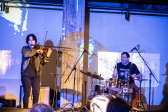 Color image of two performers on stage. The performer on the left is standing as they sing into a microphone and play the violin. The performer on the left is sitting down playing the drums with their mouth open in an expressive way. The drummer and the back wall are bathed in blue light, with blocks of white abstract patterns.
