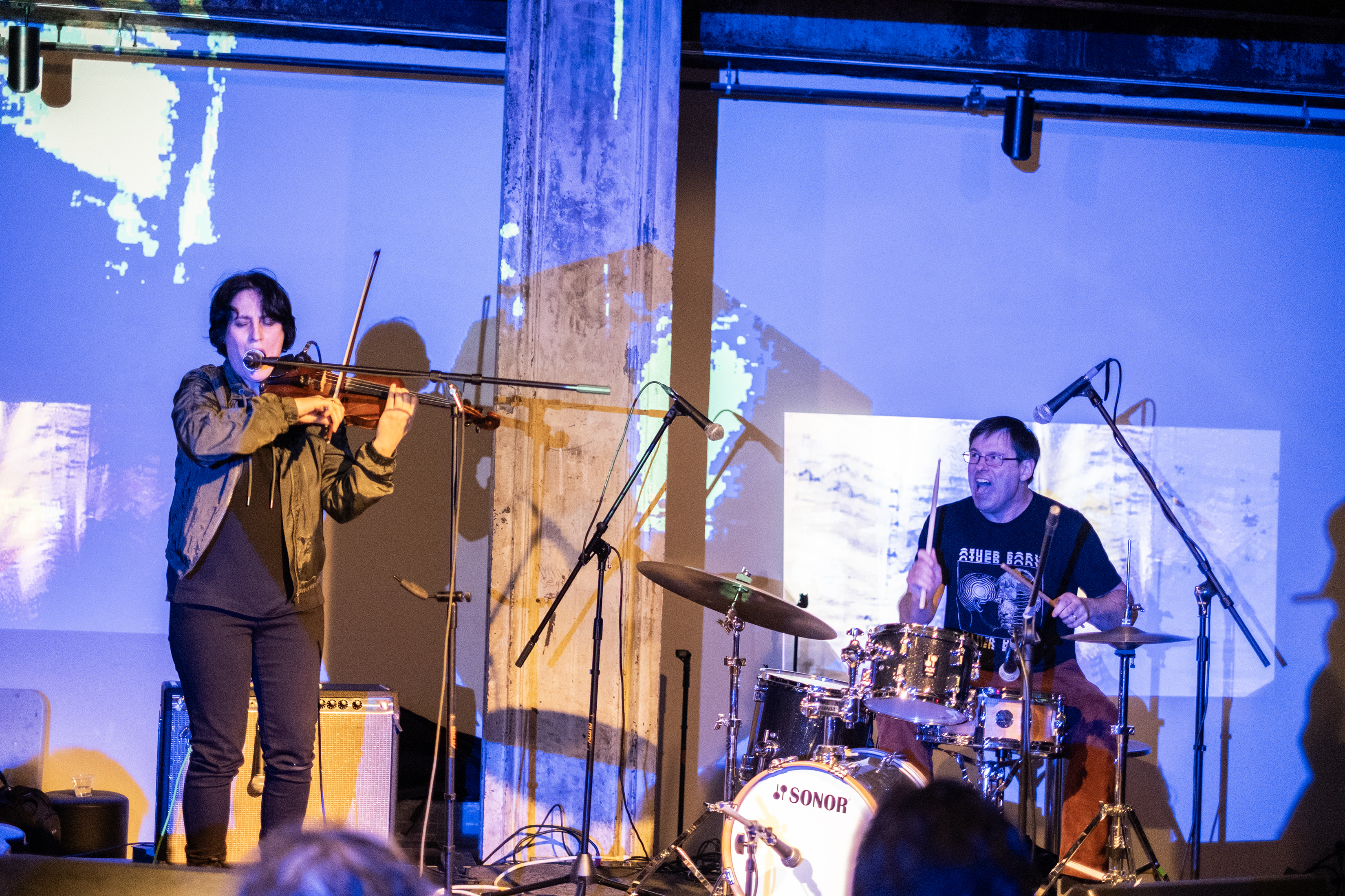 Color image of two performers on stage. The performer on the left is standing as they sing into a microphone and play the violin. The performer on the left is sitting down playing the drums with their mouth open in an expressive way. The drummer and the back wall are bathed in blue light, with blocks of white abstract patterns.
