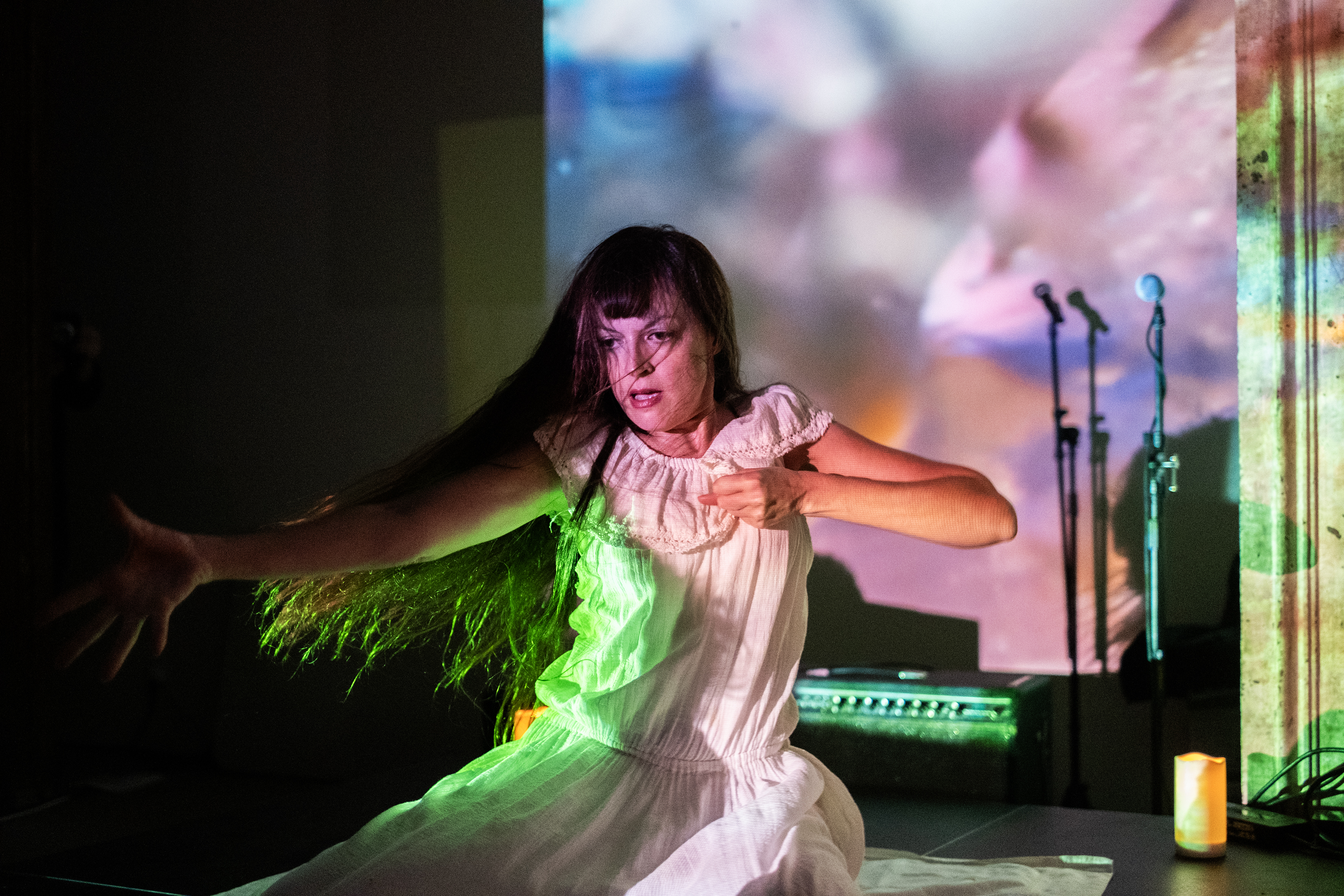Color image of a performer sitting on a stage. Their right arm is reaching out to the side with their hand open, and their left arm is bent in towards their heart with a closed fist. They are wearing a white dress. Their hair and dress are tined by a green light. The wall and column in the background are patterned with multicolored abstract projections.