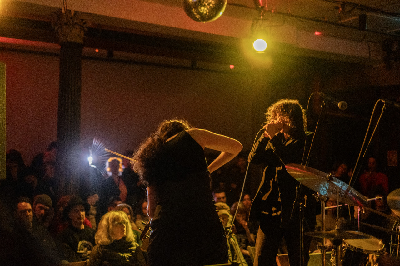 Color image of two performers, shot from behind the stage looking out to the audience. The musician on the left is bent down with their arms out and their back is facing the camera. The musician on their right is signing into a microphone. Multiple rows of audience members are facing the stage. A bright light from the back of the room creates a glare in the image. The room is dark and it is tinted yellow and pink.
