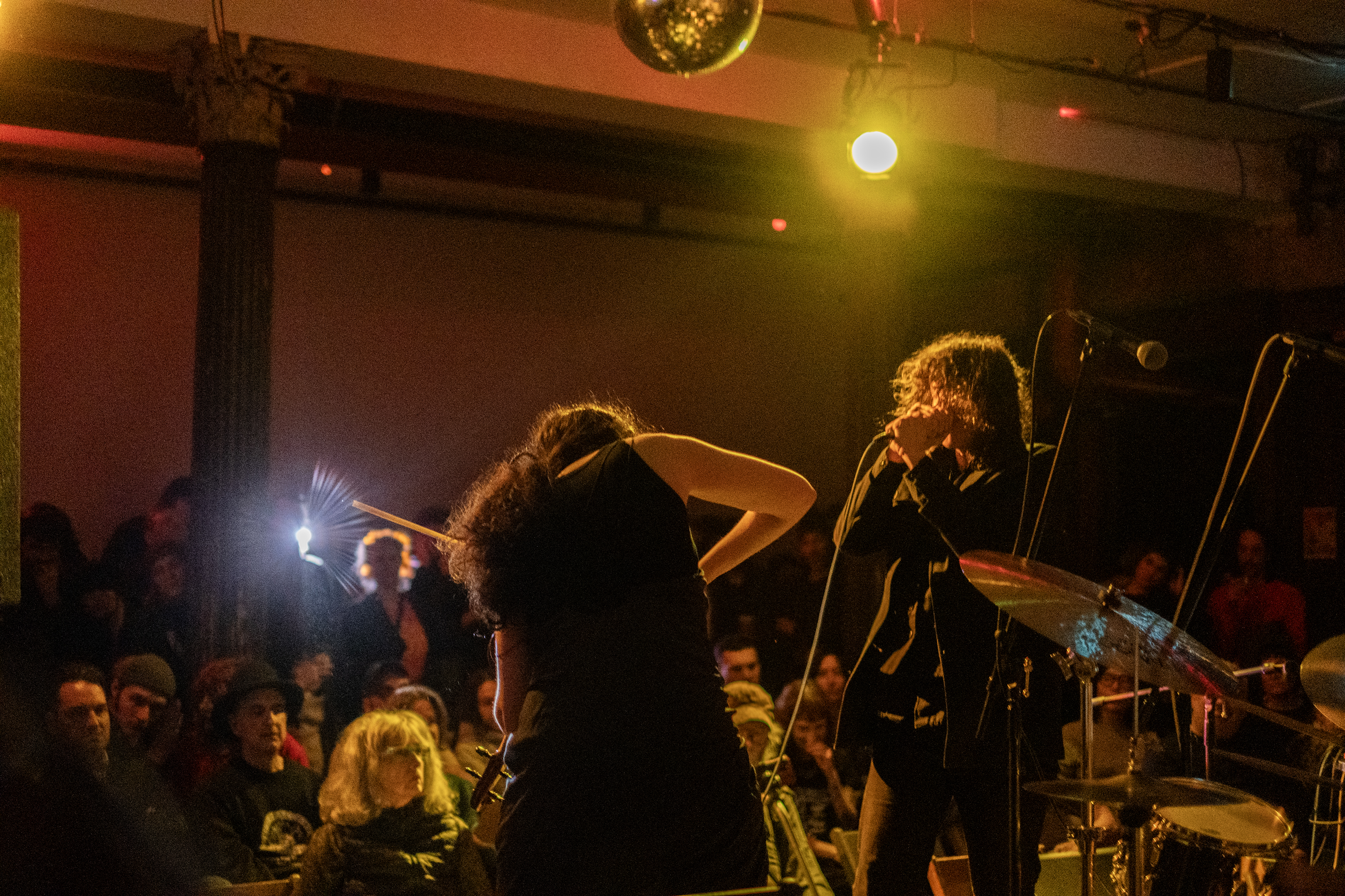 Color image of two performers, shot from behind the stage looking out to the audience. The musician on the left is bent down with their arms out and their back is facing the camera. The musician on their right is signing into a microphone. Multiple rows of audience members are facing the stage. A bright light from the back of the room creates a glare in the image. The room is dark and it is tinted yellow and pink.