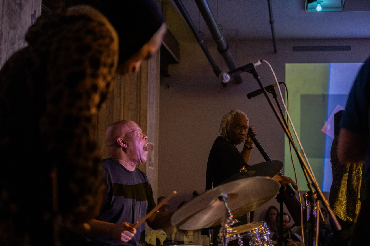 Color image of multiple performers on stage. The performer in the foreground is out of focus. In the middle one of them plays the drums, they are looking up with their mouth open. In the background, a performer faces the camera. The wall behind them is decorated with a projection of multicolored squares. A few audience members are visible in the bottom edge.