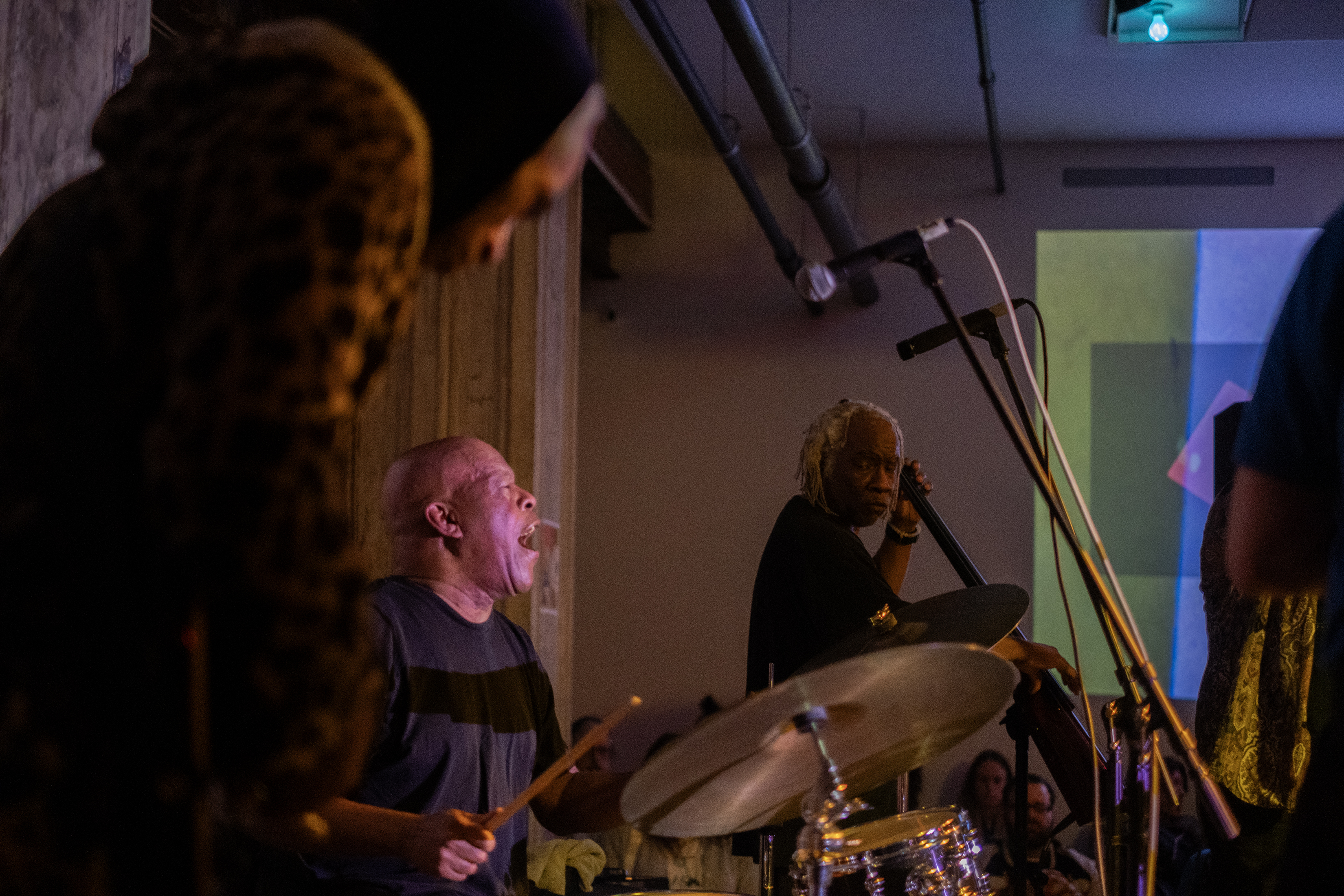 Color image of multiple performers on stage. The performer in the foreground is out of focus. In the middle one of them plays the drums, they are looking up with their mouth open. In the background, a performer faces the camera. The wall behind them is decorated with a projection of multicolored squares. A few audience members are visible in the bottom edge.