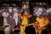 Color image of three performers on stage. In the middle a performer is standing up with their hands on their shoulders. To the right, a performer is hidden behind a large collection of tissue paper. In the back left side, a performer is sitting down in front of audio equipment. A few audience members heads are visible in the foreground. The wall behind the stage is projected on with blurry images of a crowd. 