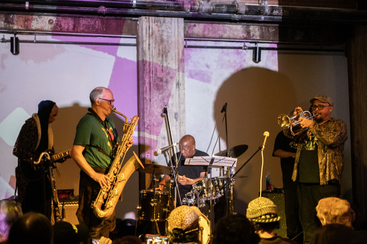 Color image of five performers on stage. From left to right, they are playing guitar, saxophone, drums, and trumpet. The room is lit up with pink and white projections. The backs of multiple audience members are visible in the bottom of the frame. A few of the performers are lit with yellow light, as well as some of the audience.