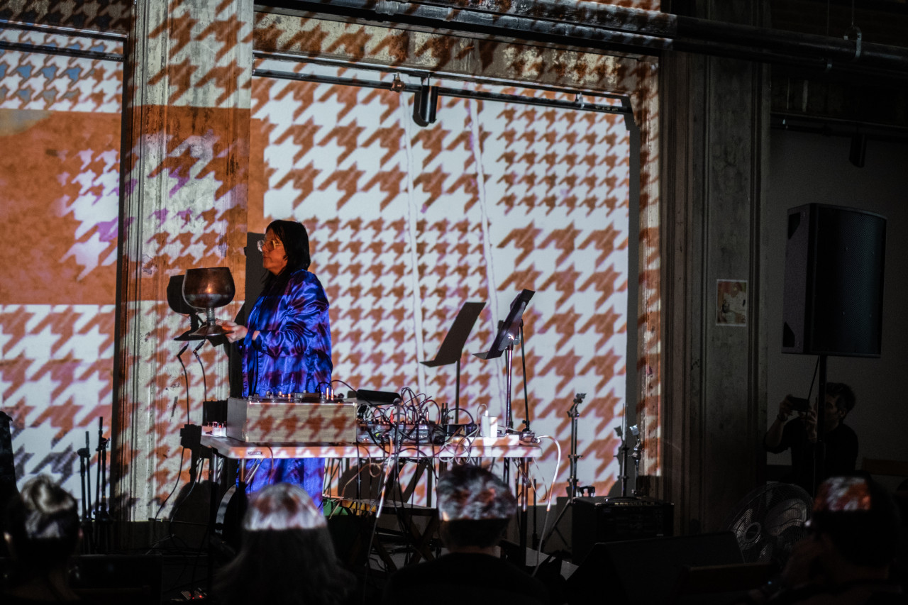 Color image of a performer on stage holding an oversized goblet. They are wearing a monochromatic purple outfit, and standing sideways to the audience. A table in front of the performer is filled with various types of electronic audio equipment. The wall behind the stage is covered in a houndstooth projected pattern.