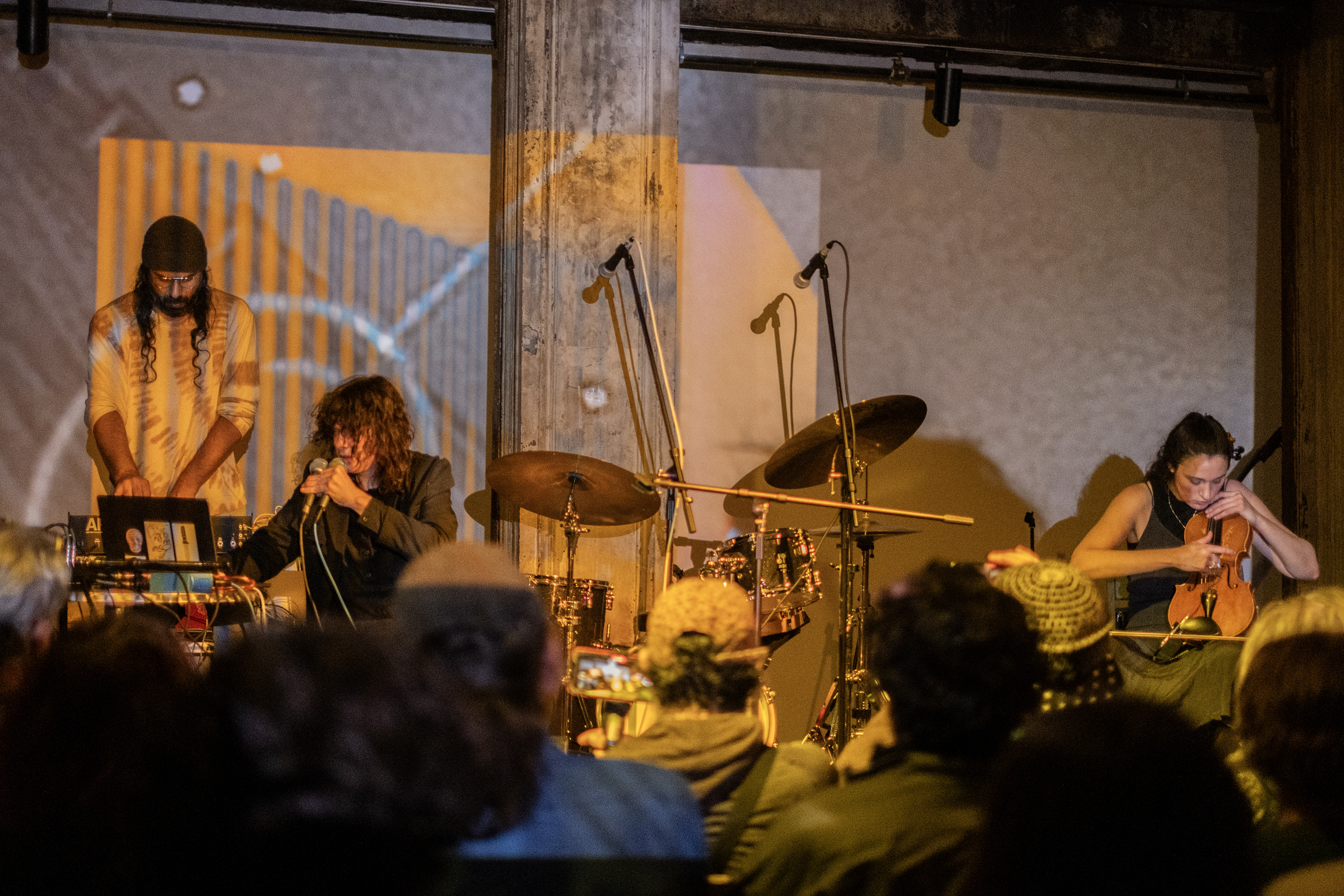 Color image of 3 performers on stage. The musician on the left is standing over electronic audio equipment while looking down. To their side a musician is sitting down holding a microphone close to their mouth. On the right side of the image a musician is sitting down playing the violin. In the middle of the stage there is a drum kit. The backs of audience members are visible in the bottom of the image. There is an abstract orange projection on the back wall, which also covers one of the musicians.
